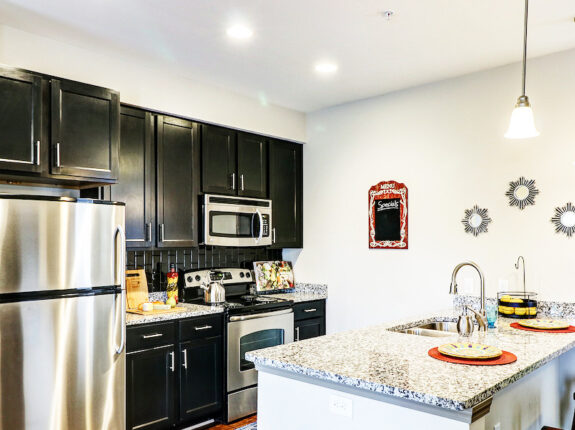 Kitchen space with chrome appliances, black cabinets, island sink with granite pattern.